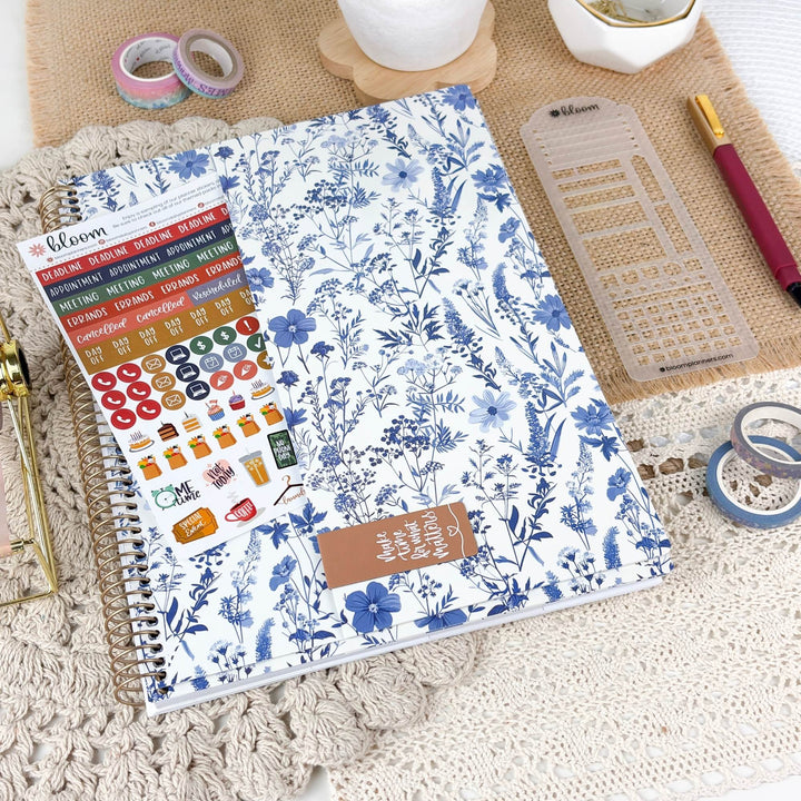 A spiral-bound planner or notebook with a blue floral print on a white background, shown in a styled flat-lay with planner stickers, washi tape, a ruler, and other stationery items.