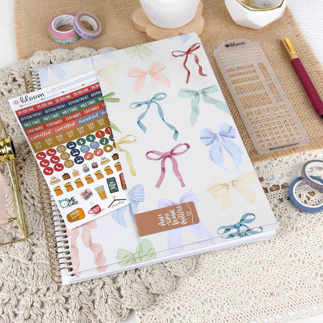 A planner or notebook cover decorated with pastel watercolor ribbon illustrations, shown in a styled flat-lay with sticker sheets, washi tape, a ruler, and other stationery items on a textured beige surface.