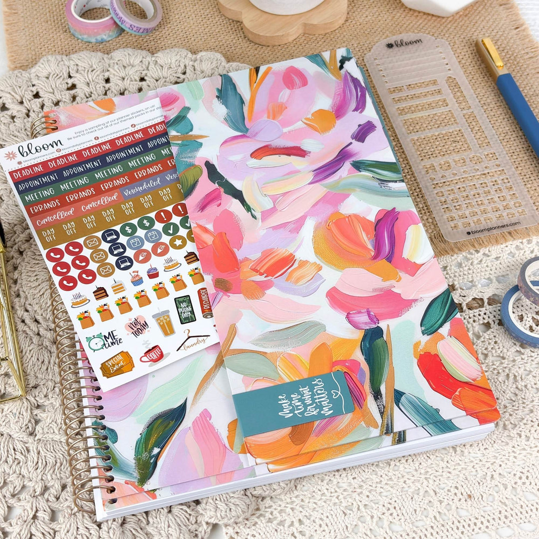 A floral planner cover or notebook with large painterly flowers in pink, orange, purple, green, and cream, styled on a crocheted surface with planner stickers, a ruler, and a pen nearby.