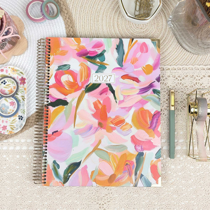 A 2027 spiral-bound planner with a colorful floral cover design, styled on a beige lace tablecloth with stationery accessories around it.