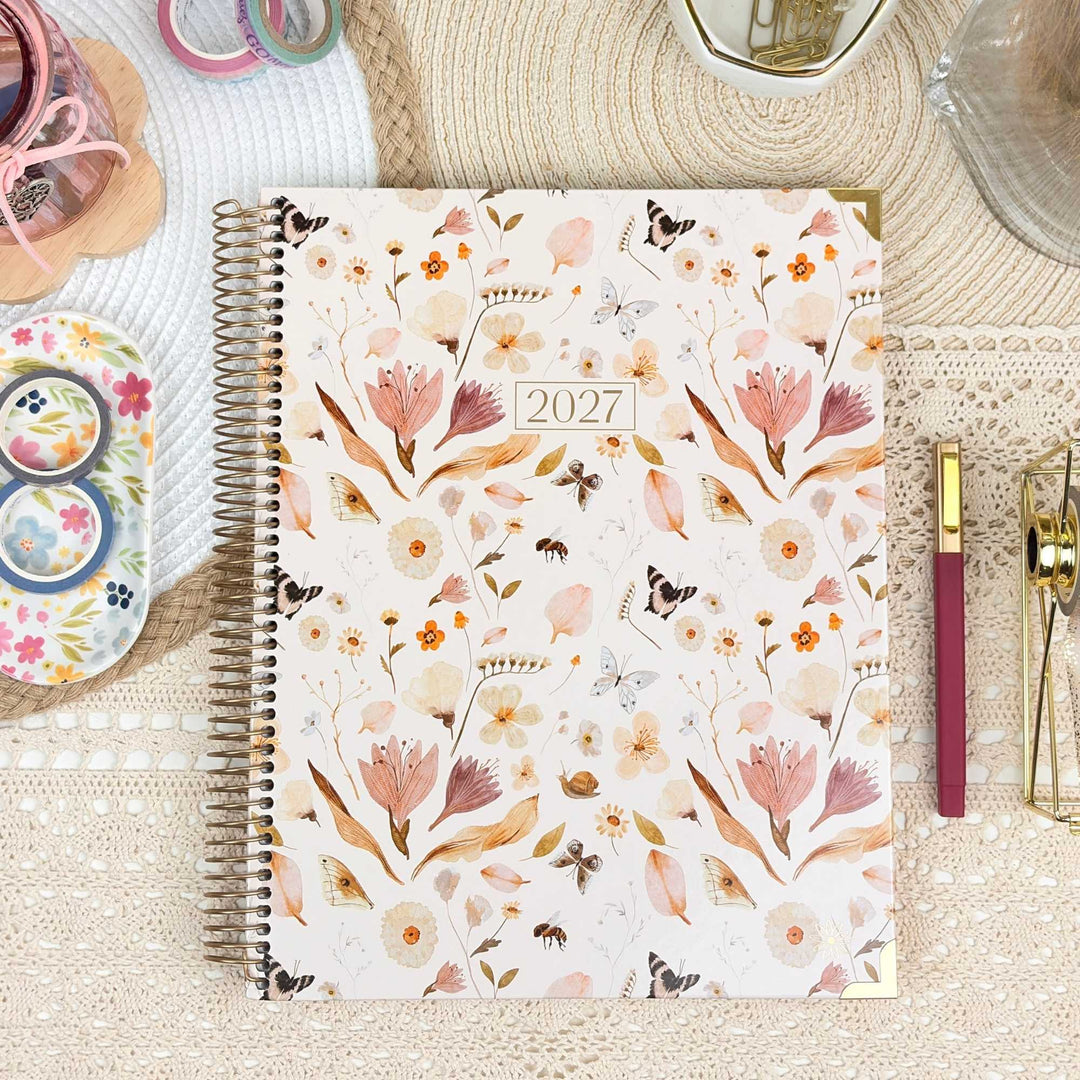 A 2027 spiral-bound planner with a floral and butterfly pattern on the cover, styled on a textured beige lace tablecloth with stationery props.