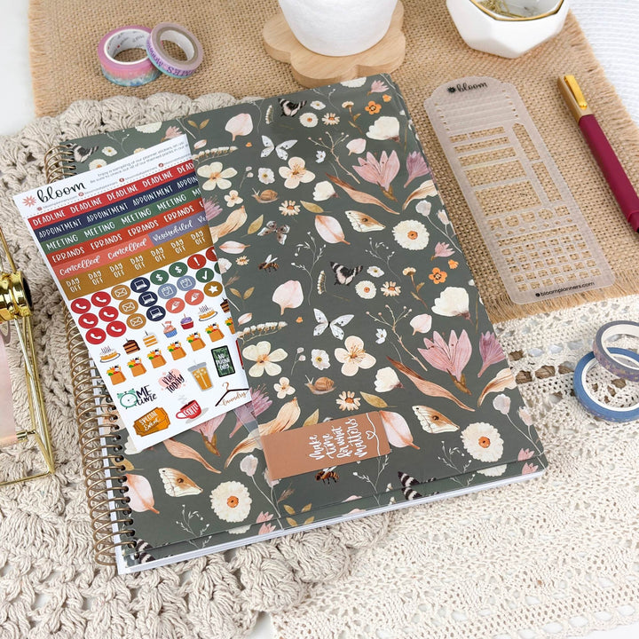 A spiral-bound planner or notebook with a dark gray floral cover is styled on a textured cream rug with planner accessories around it, including washi tape, a ruler, a pen, and sticker sheets. The brand name 'bloom' is visible on the sticker sheet and a small item in the top right.