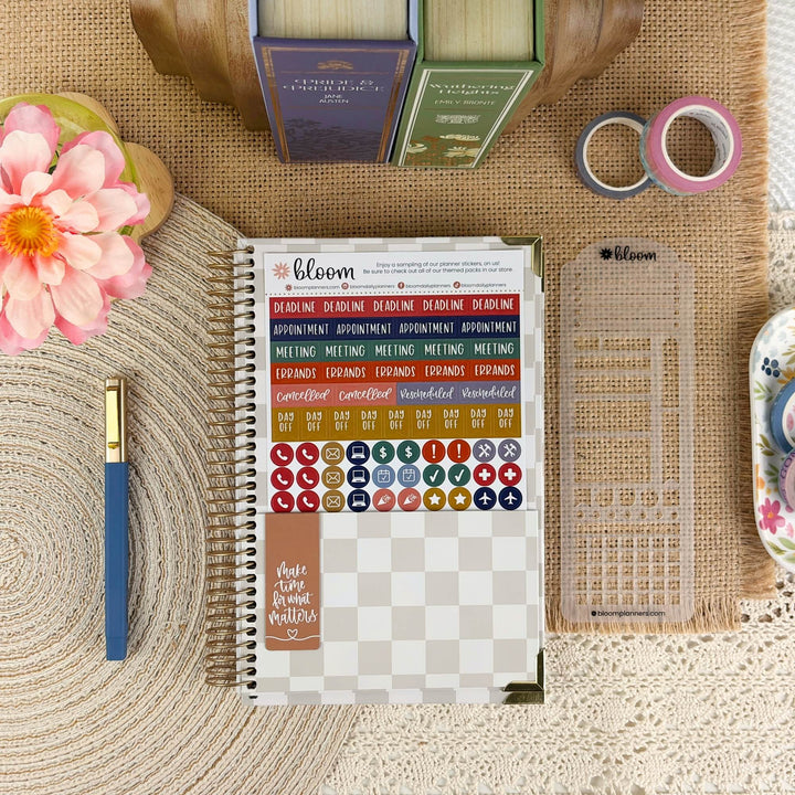 A spiral-bound planner sticker sheet set by bloom planner, displayed in a styled flat-lay on a burlap mat with a blue pen, pink flower, washi tape, and other stationery accessories.