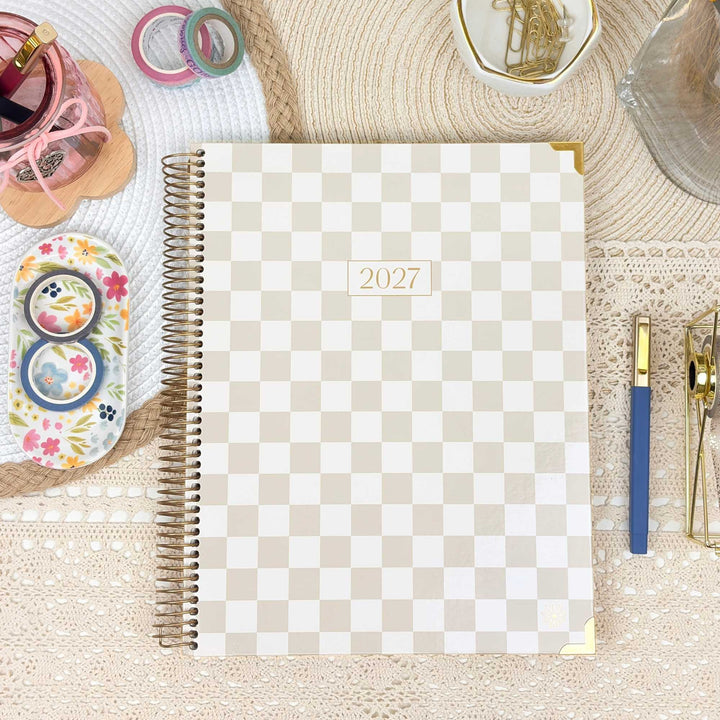 A 2027 spiral-bound planner with a beige and white checkerboard pattern, styled in a flat-lay arrangement on a lace tablecloth with stationery accessories.