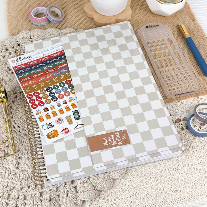 A spiral-bound planner or notebook with a light gray and white checkerboard cover lies on a textured cream knit surface. A sheet of colorful planner stickers is placed on top, along with a clear plastic ruler, a blue pen, and other stationery items.