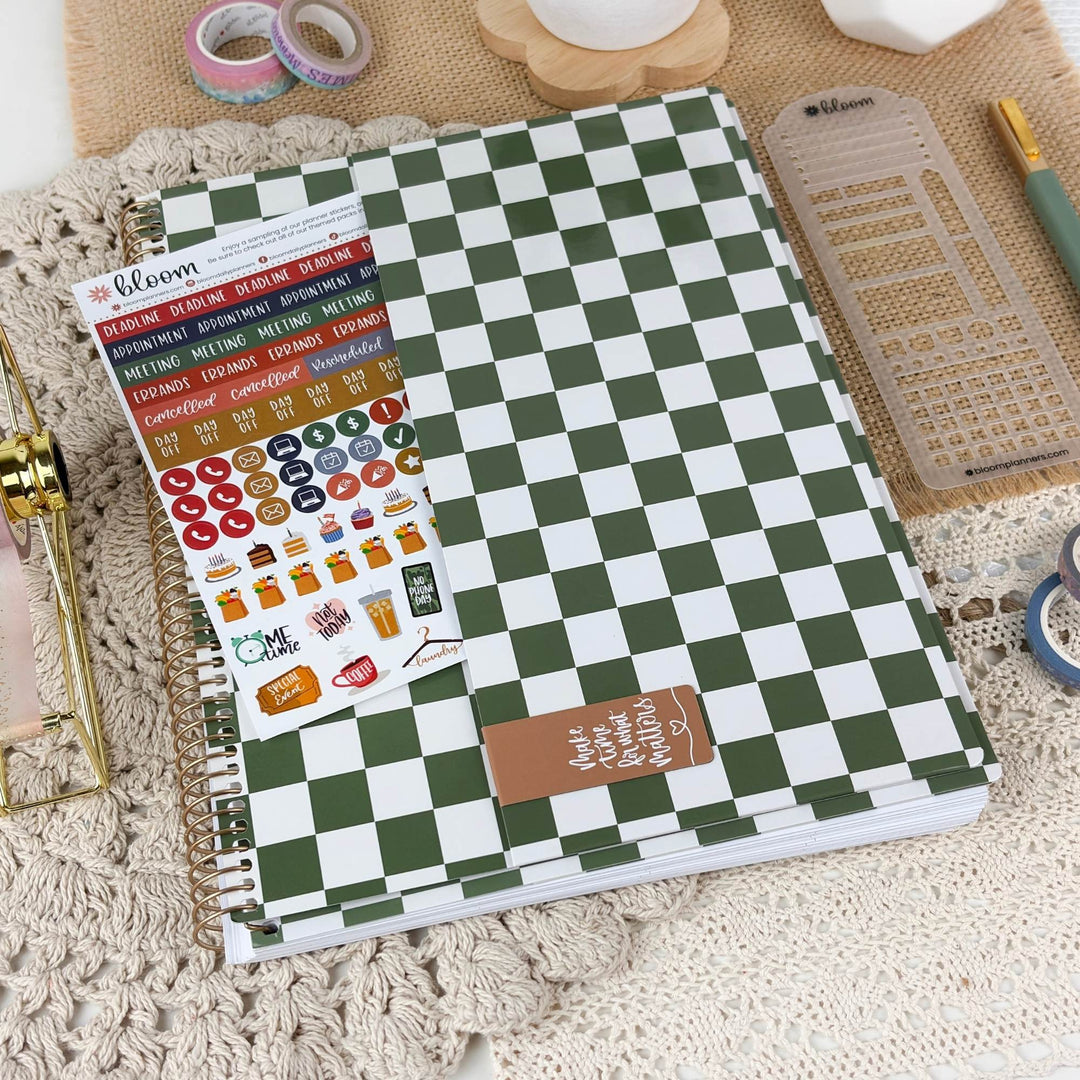 A spiral-bound planner or notebook with a green and white checkerboard cover lies on a textured cream lace surface. A sheet of colorful planner stickers is placed on top, along with a small brown tag.