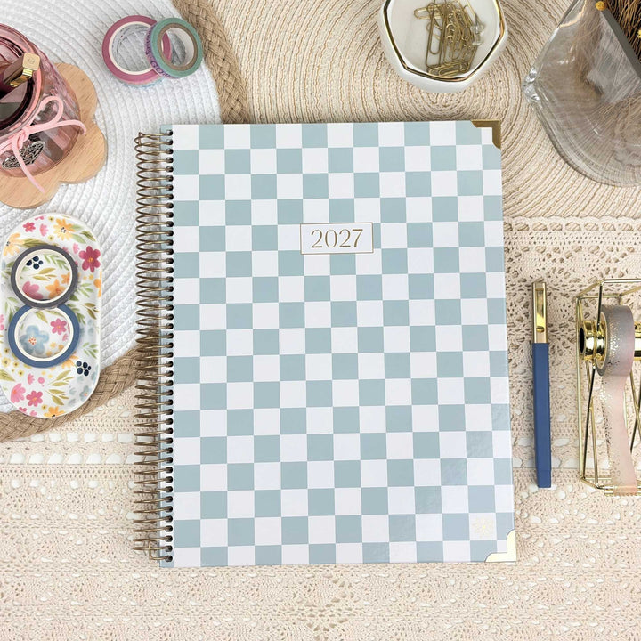 A spiral-bound 2027 planner with a light blue and white checkerboard cover, styled in a flat lay on a beige lace tablecloth with stationery accessories.