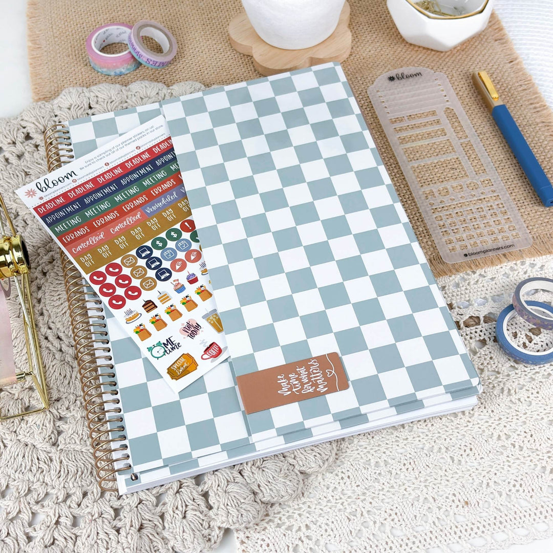 A spiral-bound planner or notebook with a light blue and white checkerboard cover is shown on a textured beige surface, alongside sticker sheets, washi tape, a ruler, and other stationery accessories.