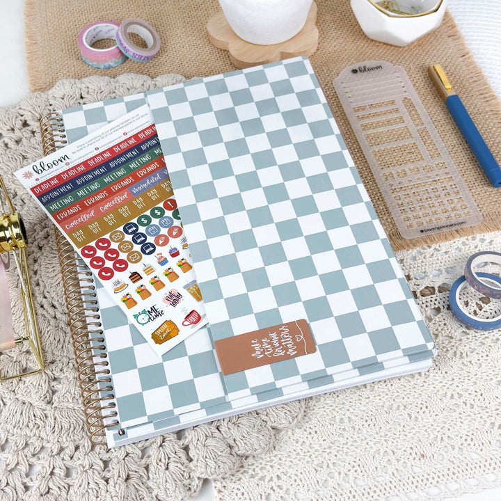 A spiral-bound planner or notebook with a light blue and white checkerboard cover is shown on a textured beige surface, alongside sticker sheets, washi tape, a ruler, and other stationery accessories.