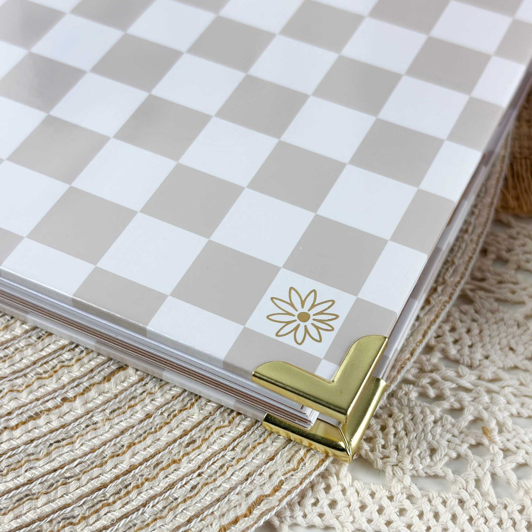 A close-up of a planner or notebook cover with a beige and white checkerboard pattern, featuring a small gold daisy logo and a gold metal corner accent, resting on a cream crocheted textile.