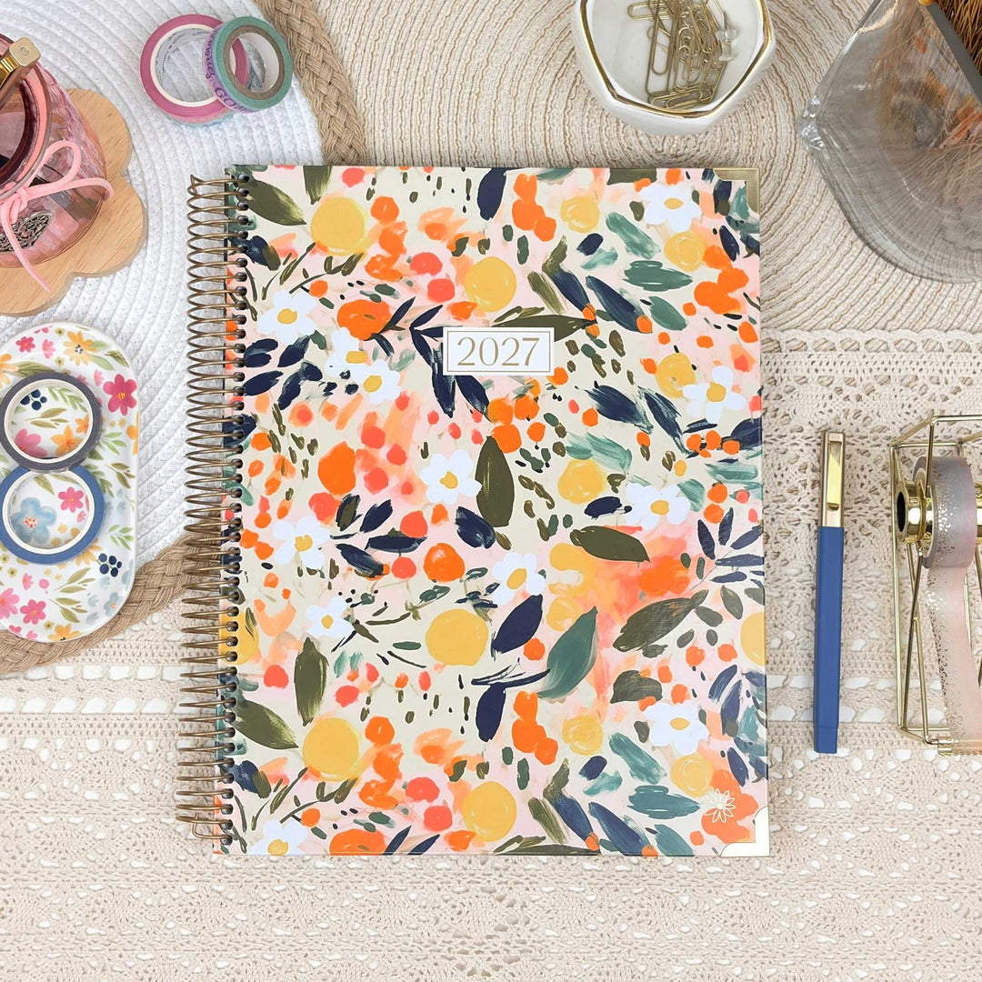 A spiral-bound 2027 planner with a colorful floral and citrus watercolor cover pattern, styled in a flat-lay arrangement on a beige lace tablecloth with stationery accessories.