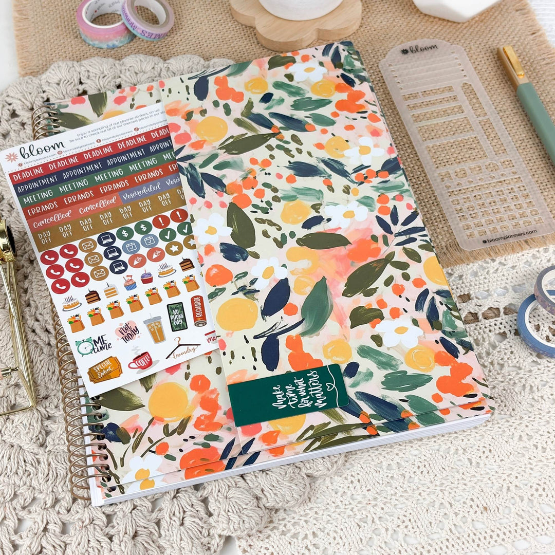 A floral planner cover or notebook with a colorful watercolor-style leaf and fruit pattern, shown alongside sticker sheets, a clear plastic divider, washi tape, and a pen on a textured neutral background.