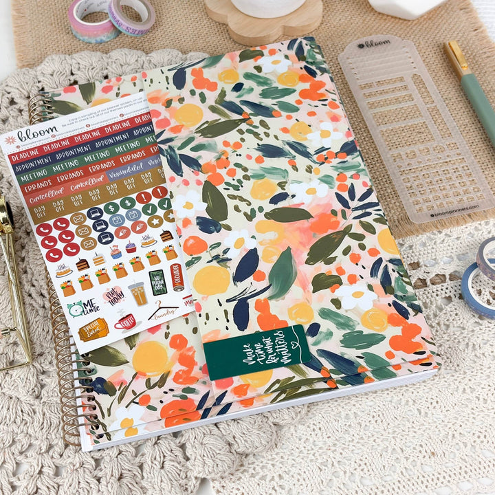 A floral planner cover or notebook with a colorful watercolor-style leaf and fruit pattern, shown alongside sticker sheets, a clear plastic divider, washi tape, and a pen on a textured neutral background.