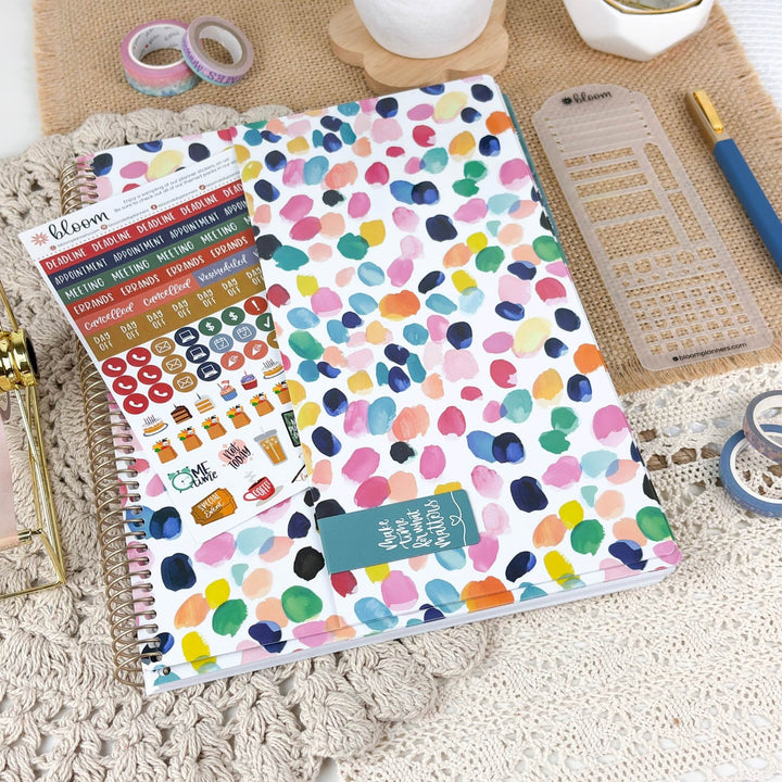 A spiral-bound planner or notebook with a white cover covered in colorful paint-drip patterns, styled on a textured beige surface with stationery accessories including washi tape, a clear ruler, a blue pen, and sticker sheets. The Bloom brand name is visible on the sticker sheet.