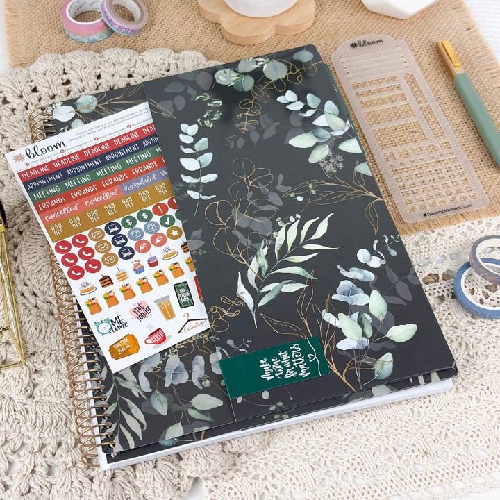 A floral planner or notebook with a black cover, green and white botanical leaves, and gold vine details, shown on a beige textured surface with planner accessories including stickers, a pen, and a clear ruler.