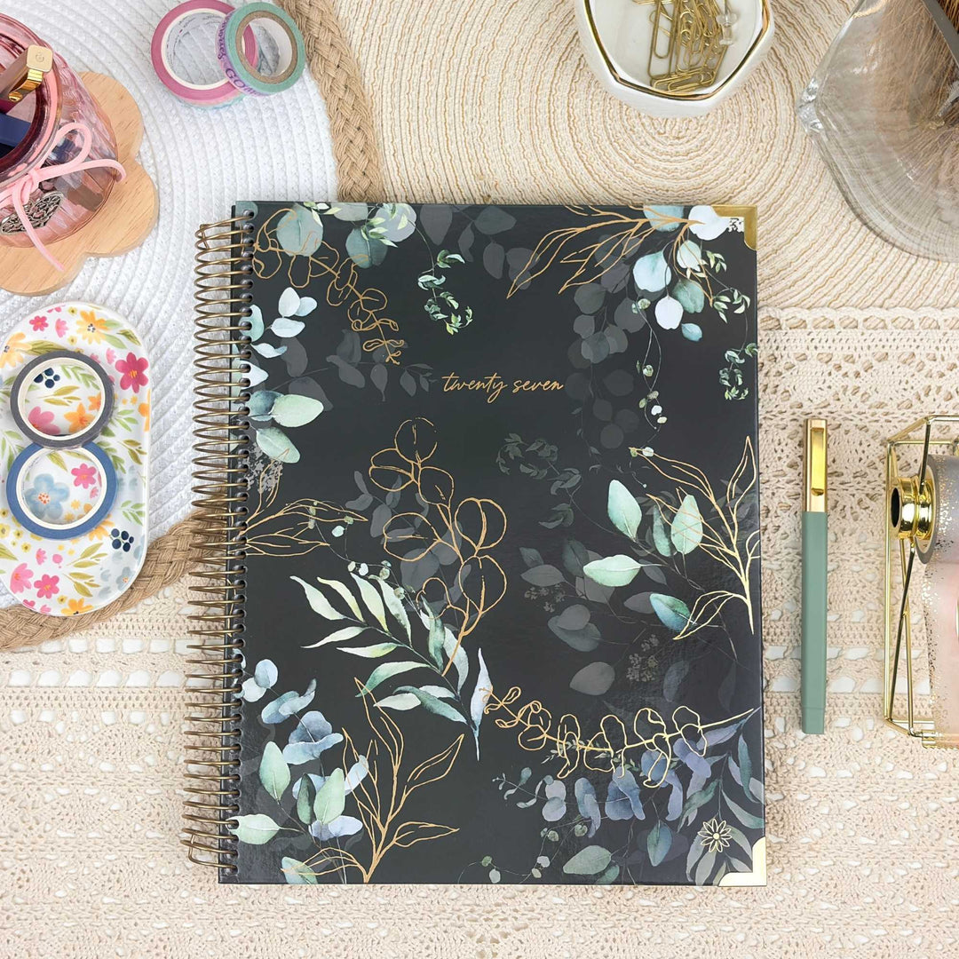 A spiral-bound planner or notebook with a black cover decorated with watercolor-style green and blue botanical leaves and gold foil details, placed on a beige lace surface with stationery props around it.