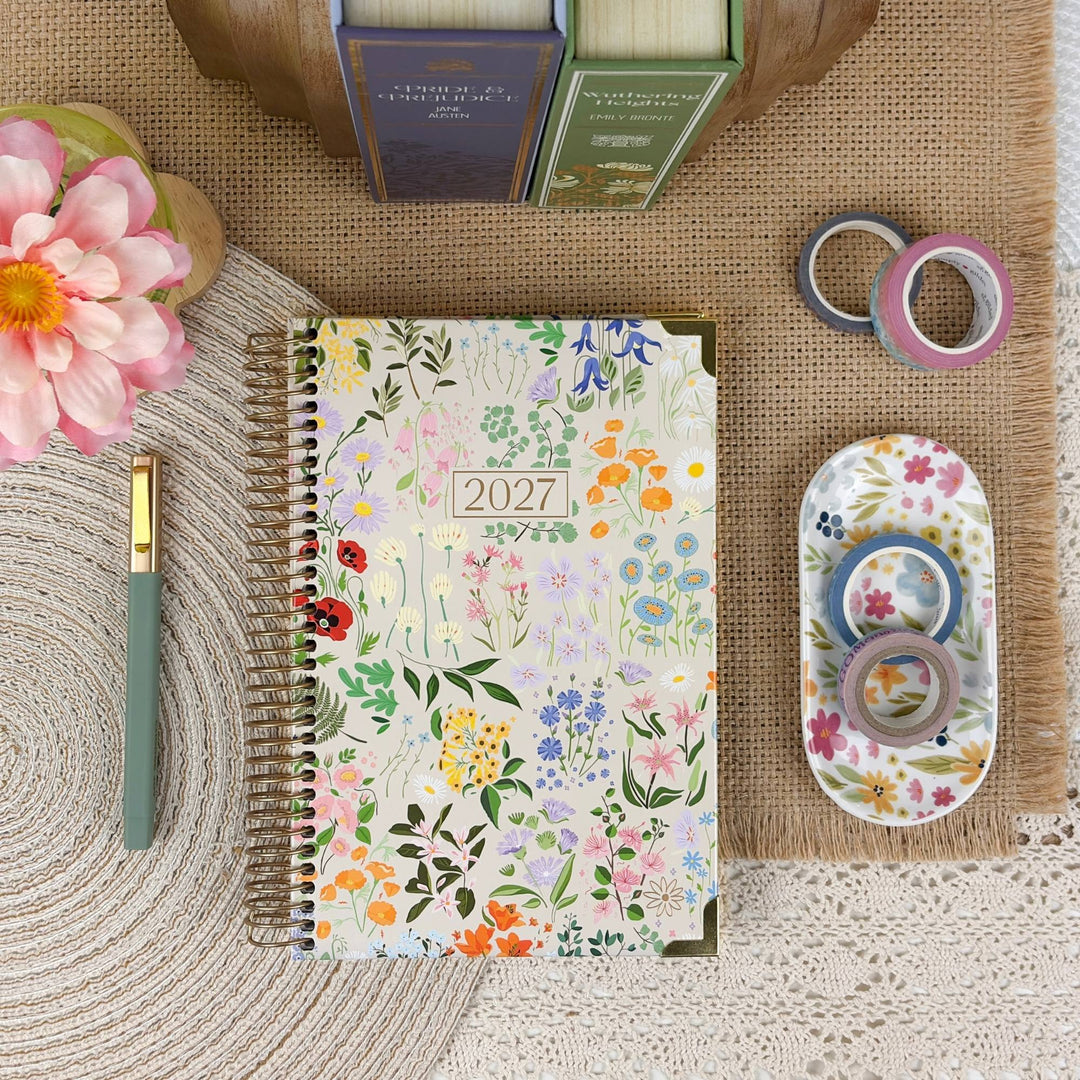 A floral 2027 spiral-bound planner is displayed in a styled flat-lay arrangement on a burlap mat, surrounded by a green pen, washi tapes, a small floral dish, books, and a pink flower.