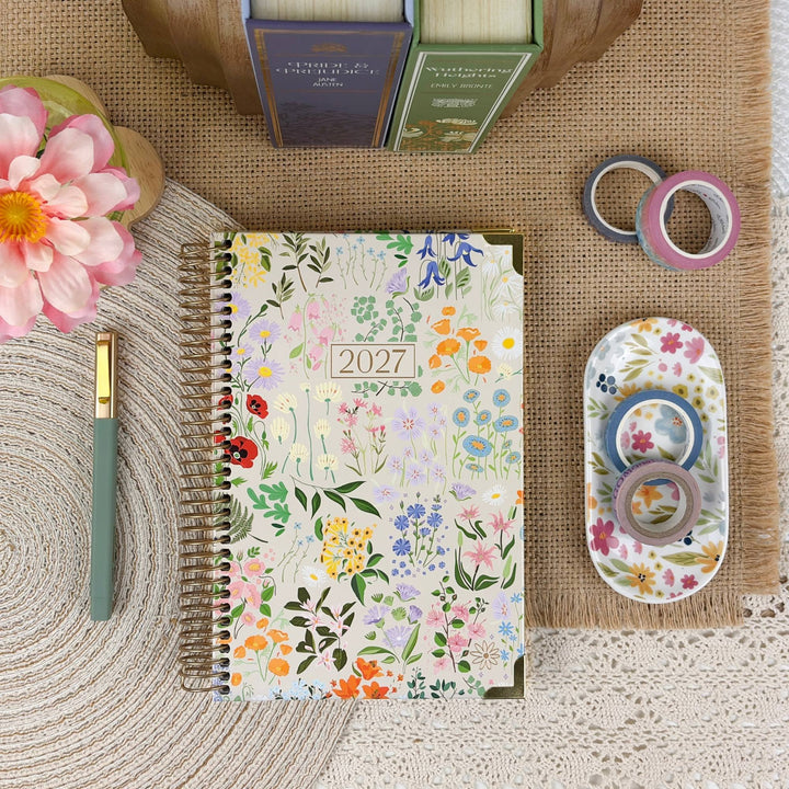 A floral 2027 spiral-bound planner is displayed in a styled flat-lay arrangement on a burlap mat, surrounded by a green pen, washi tapes, a small floral dish, books, and a pink flower.