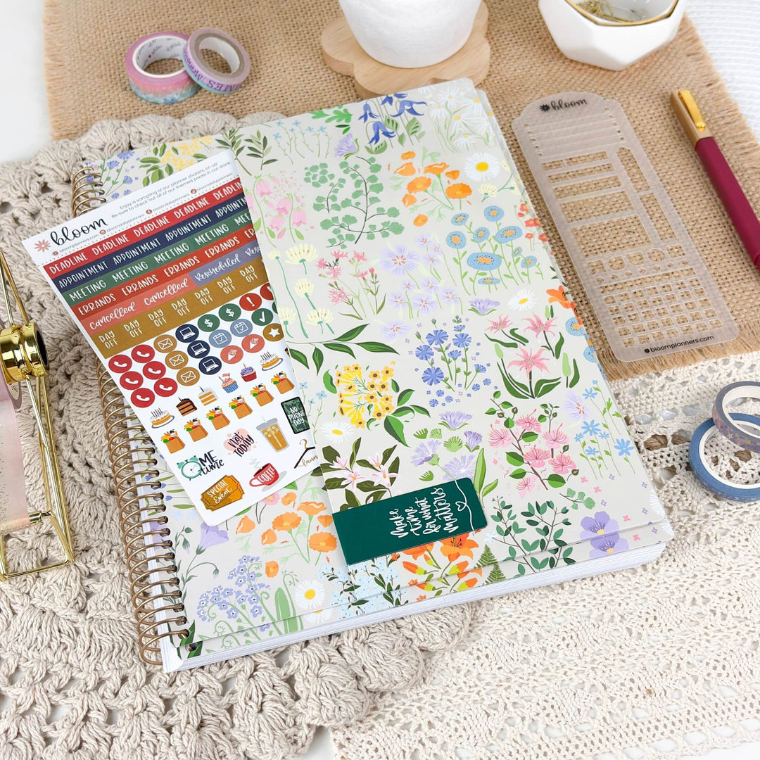 A floral planner sticker sheet and matching planner stickers are displayed on a spiral-bound planner, surrounded by desk accessories like washi tapes, a ruler, and a pen on a neutral fabric background.