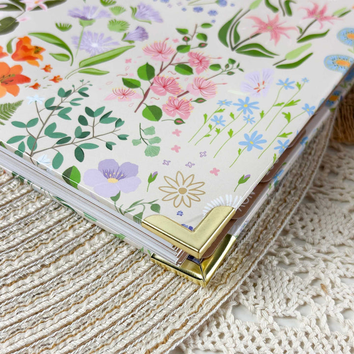 A close-up of a floral-patterned notebook or planner cover with a gold metal corner clasp, resting on a cream lace textile background.