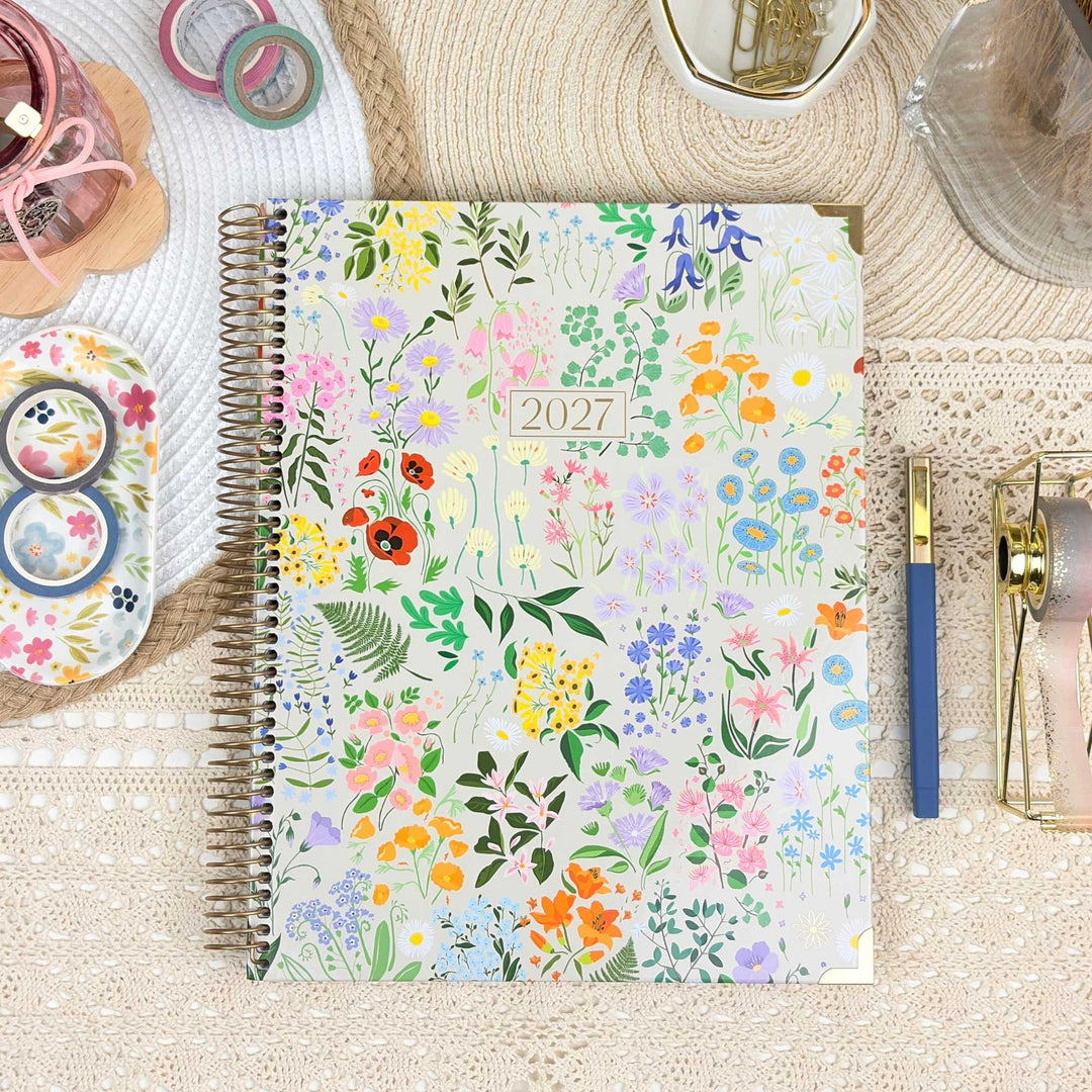 A spiral-bound 2027 planner with a colorful wildflower pattern on the cover, styled on a beige lace surface with stationery props including a blue pen, washi tape, and a glass bowl.