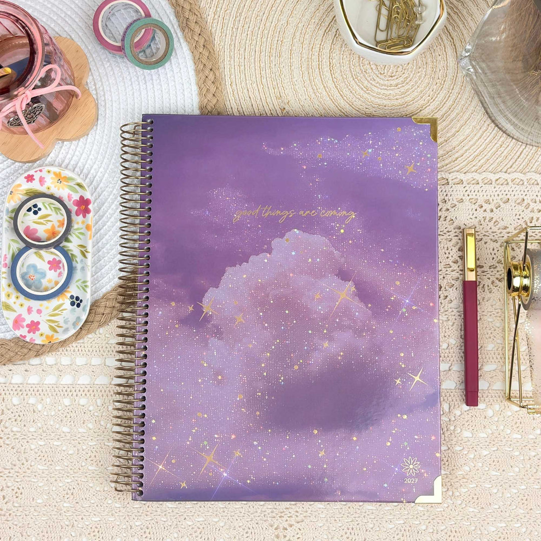 A spiral-bound planner or notebook with a purple and lavender galaxy cloud cover, styled in a flat lay with stationery accessories on a lace tablecloth.