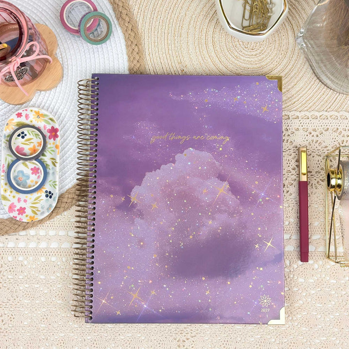 A spiral-bound planner or notebook with a purple and lavender galaxy cloud cover, styled in a flat lay with stationery accessories on a lace tablecloth.