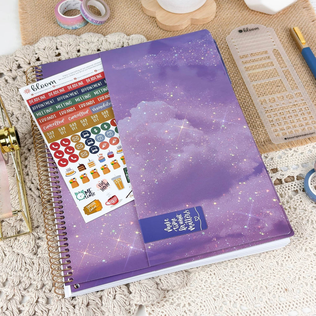 A spiral-bound planner or notebook with a purple galaxy cover is styled on a lace surface with desk accessories, including washi tape, a ruler, and sticker sheets. The cover has a purple cloudy space design with white stars and a blue label reading 'Make it worth remembering'.