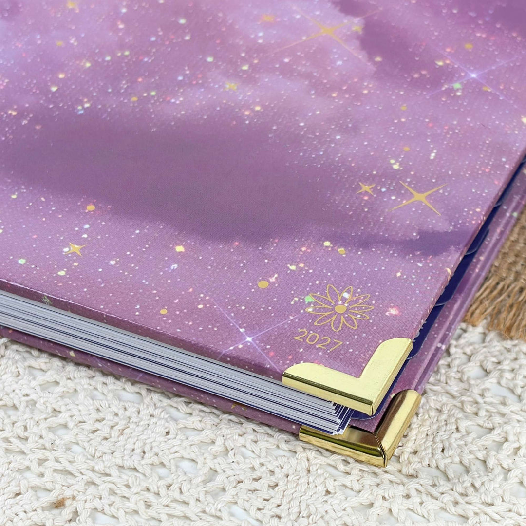 A close-up of a purple glittery notebook or planner cover with gold star and flower details, showing the year 2027 and gold corner protectors on a textured cream surface.
