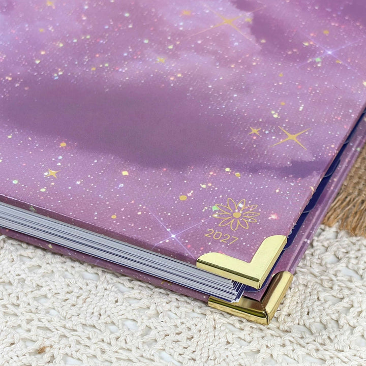 A close-up of a purple glittery notebook or planner cover with gold star and flower details, showing the year 2027 and gold corner protectors on a textured cream surface.
