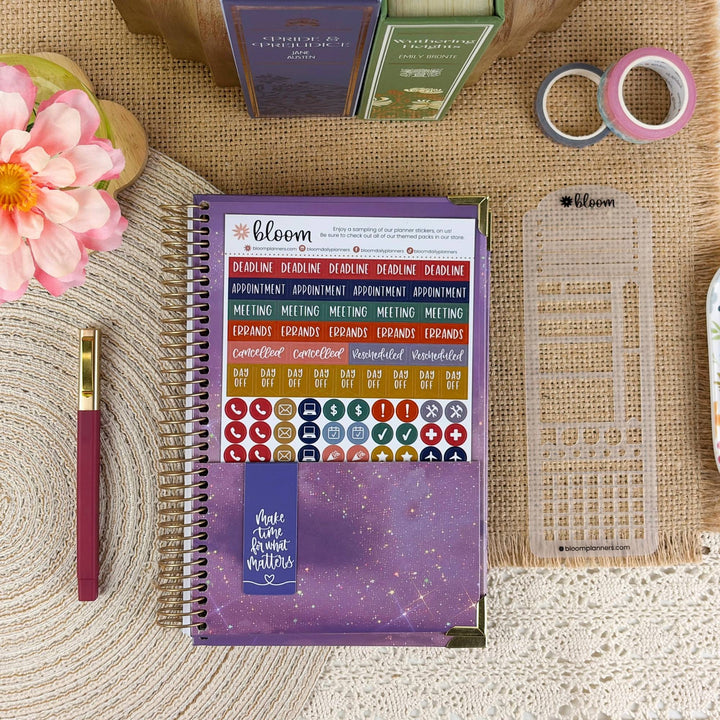 A spiral-bound planner or notebook with colorful sticker sheets and a purple cover, styled on a burlap surface with a pink flower, pen, washi tape, and other stationery items nearby.