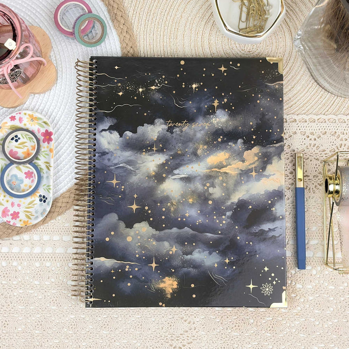 A spiral-bound notebook with a celestial cloud cover design in navy, gray, and gold, styled on a beige lace surface with stationery props.