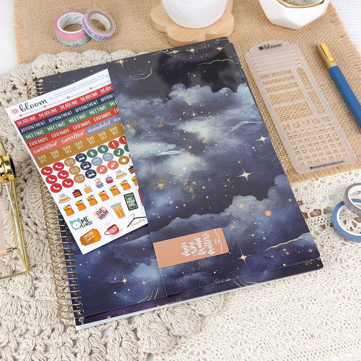 A spiral-bound planner or notebook with a dark blue and black galaxy night-sky cover design, styled on a beige textured surface with planner stickers, washi tape, a ruler, and a pen nearby.