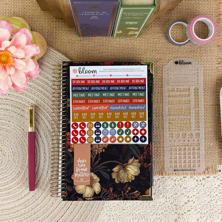 A spiral-bound planner or notebook with a floral cover and a sticker sheet attached, styled on a burlap surface with a pink flower, a pen, washi tape, and other stationery items around it. The visible brand on the product is bloom.