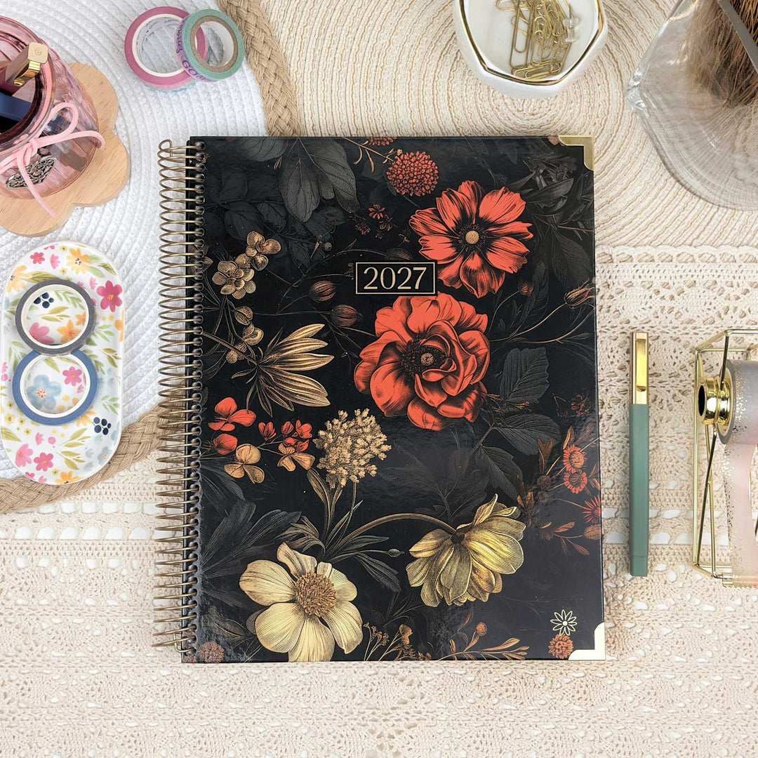 A spiral-bound 2027 planner with a dark floral cover design, styled on a beige lace surface with desk accessories including a green pen, washi tape, and a small tray.