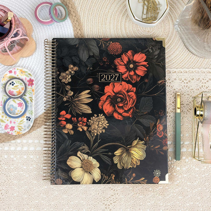 A spiral-bound 2027 planner with a dark floral cover design, styled on a beige lace surface with desk accessories including a green pen, washi tape, and a small tray.