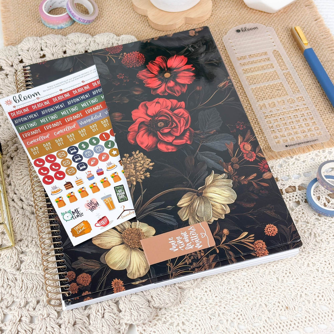 A floral planner or notebook with a black cover featuring red and cream flowers, shown with a sheet of colorful planner stickers and surrounded by stationery on a textured beige surface.