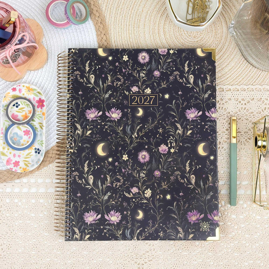 A 2027 spiral-bound planner with a dark navy floral and celestial pattern, styled on a beige lace surface with stationery props.