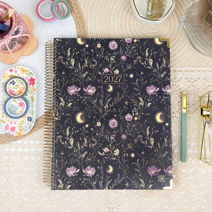 A 2027 spiral-bound planner with a dark navy floral and celestial pattern, styled on a beige lace surface with stationery props.