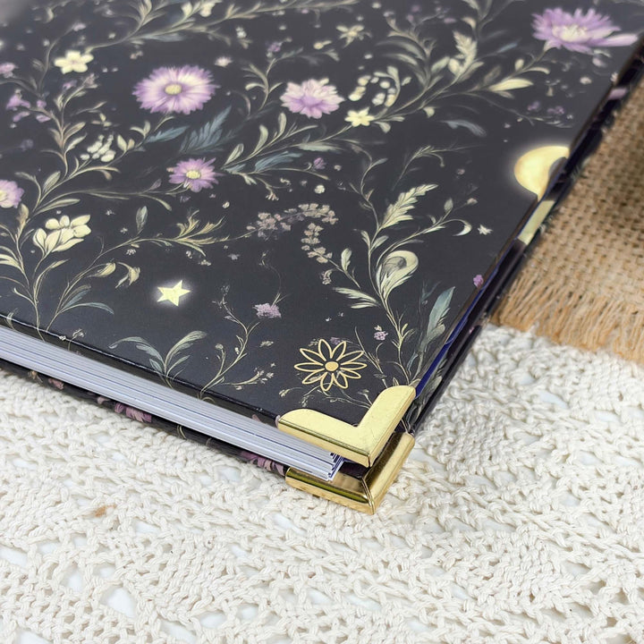 A close-up of a decorative notebook or journal with a black cover featuring a floral and celestial print in lavender, gold, and cream. The notebook is styled on a white lace surface next to a piece of burlap fabric.