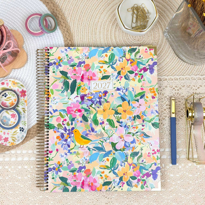 A 2027 spiral-bound planner with a colorful floral and bird pattern, styled in a flat-lay desk setting with stationery accessories.