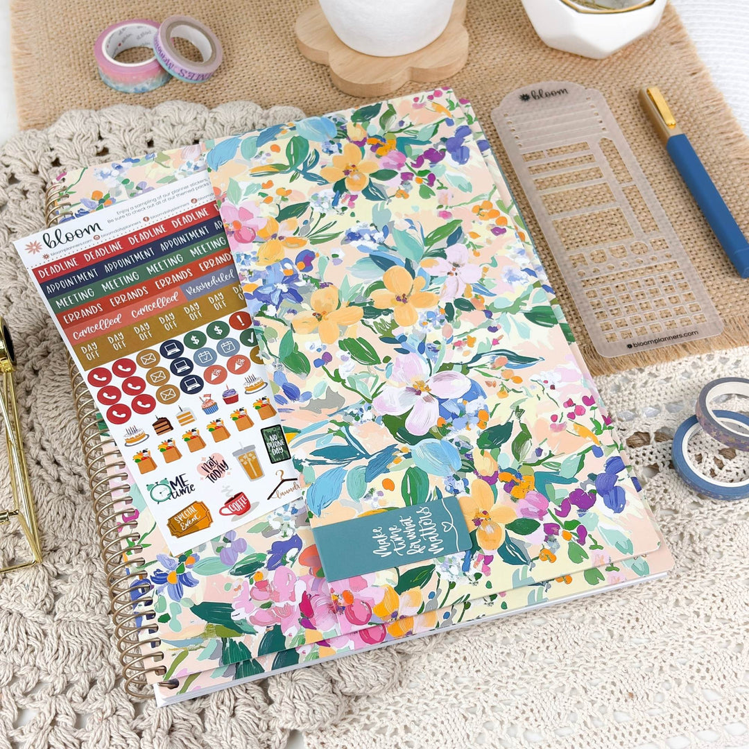 A spiral-bound planner or notebook with a colorful floral cover lies on a textured cream surface, surrounded by stationery items including washi tapes, a ruler, a pen, and sticker sheets. The cover features a pastel watercolor floral pattern with pink, yellow, blue, green, and purple flowers.