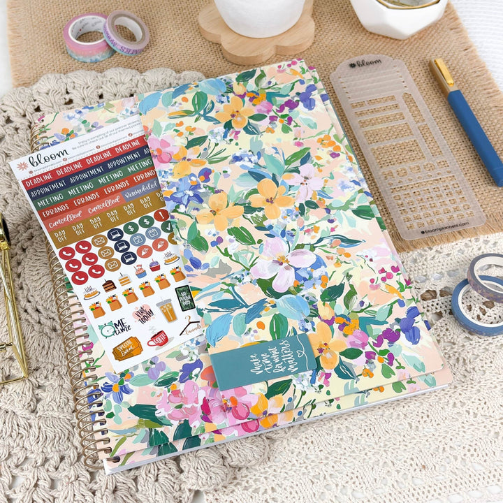 A spiral-bound planner or notebook with a colorful floral cover lies on a textured cream surface, surrounded by stationery items including washi tapes, a ruler, a pen, and sticker sheets. The cover features a pastel watercolor floral pattern with pink, yellow, blue, green, and purple flowers.