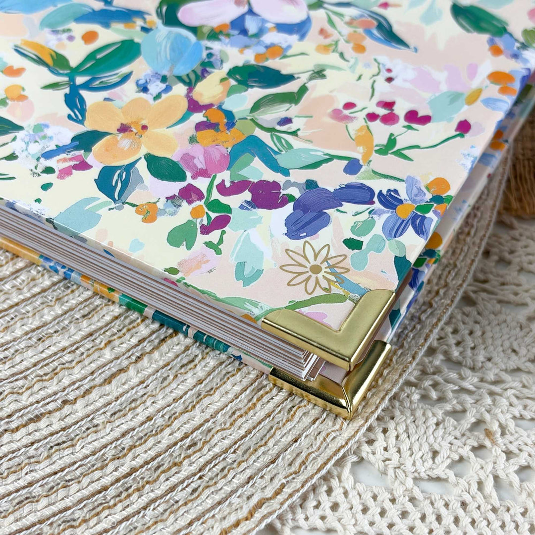 A close-up of a floral-patterned notebook or journal with a cream cover, gold metal corner protectors, and a stack of pages, resting on a textured cream woven textile.