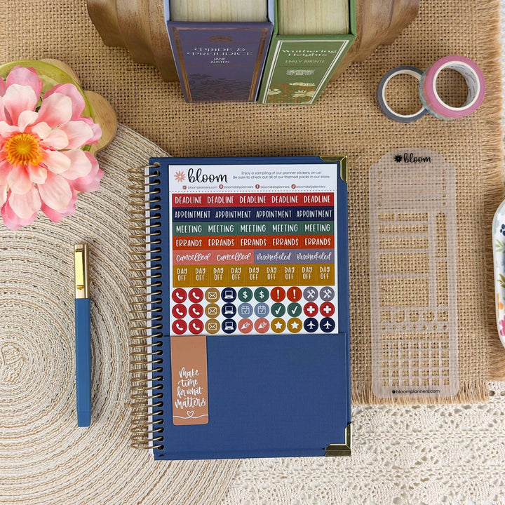 A blue spiral-bound planner or notebook with decorative stickers on the cover, arranged on a woven beige surface with a pink flower, pen, washi tape, books, and a clear grid template nearby.