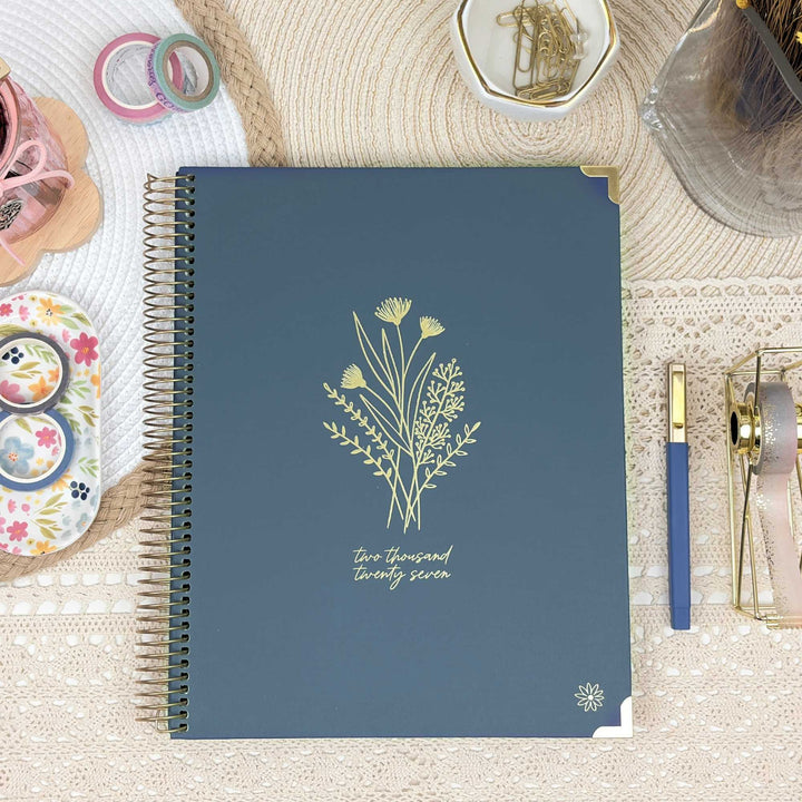 A navy blue spiral-bound planner or notebook with gold floral line art and the text 'Two thousand twenty seven' on the cover, styled on a beige textured surface with pens, washi tape, and small decorative items around it.
