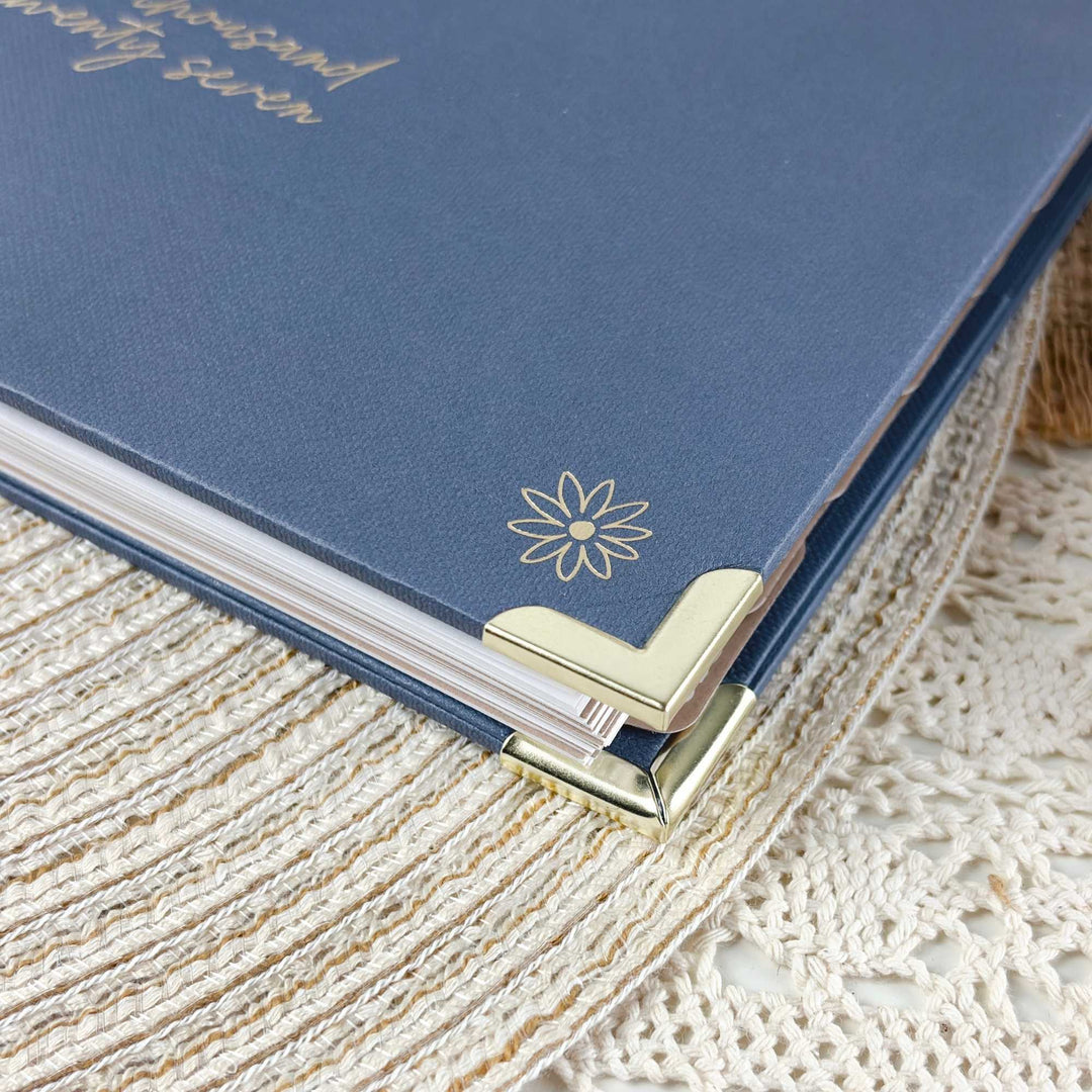 A close-up of a blue hardcover journal or notebook with gold foil script and a flower emblem, resting on a cream lace textile with gold corner protectors visible.