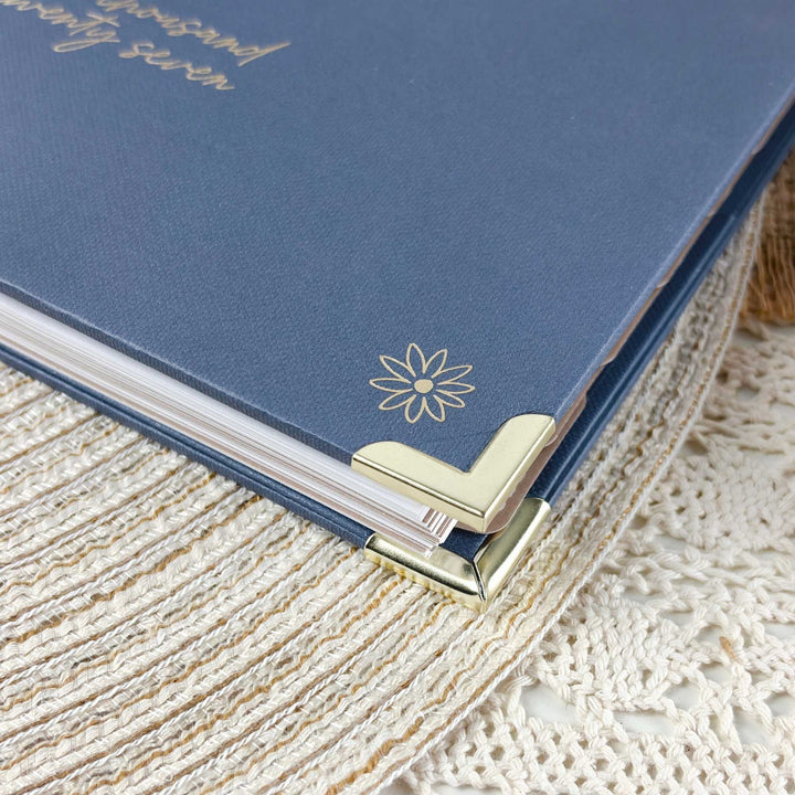 A close-up of a blue hardcover journal or notebook with gold foil script and a flower emblem, resting on a cream lace textile with gold corner protectors visible.