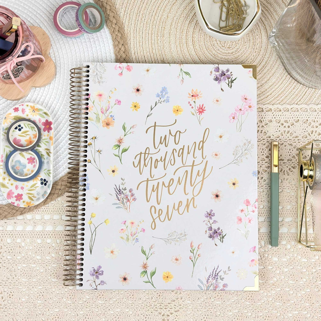 A spiral-bound planner or journal with a white cover decorated with delicate watercolor-style flowers and gold script reading 'two thousand twenty seven,' styled on a woven mat with stationery accessories around it.
