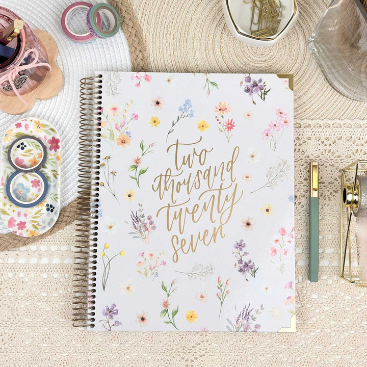 A spiral-bound planner or journal with a white cover decorated with delicate watercolor-style flowers and gold script reading 'two thousand twenty seven,' styled on a woven mat with stationery accessories around it.